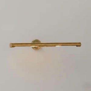 May include: A brass-toned picture light fixture. The fixture has a long, horizontal bar with two small lights and a circular base. The light is mounted on a neutral-colored wall.