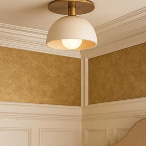 May include: A modern ceiling light fixture with a white, dome-shaped shade and a brass-colored base. The light has a round, glowing bulb. The fixture is mounted on a white ceiling with decorative molding.