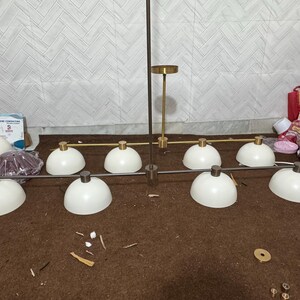 May include: A linear chandelier featuring eight white dome-shaped shades arranged in two rows. The fixture has a central vertical rod and horizontal arms finished in a metallic gold tone, with matching gold-toned hardware connecting each shade.