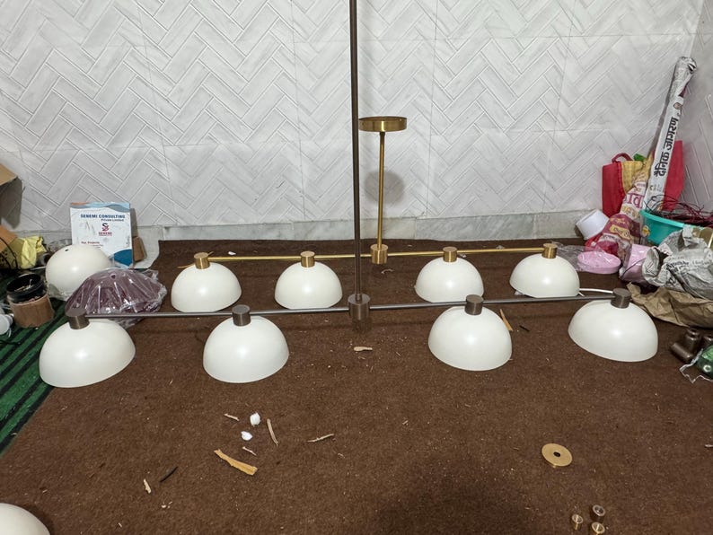 May include: A linear chandelier featuring eight white dome-shaped shades arranged in two rows. The fixture has a central vertical rod and horizontal arms finished in a metallic gold tone, with matching gold-toned hardware connecting each shade.