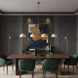 May include: A rectangular wooden dining table with four dark green upholstered chairs, a linear pendant light with four cream-colored dome shades, and two thin floor lamps positioned in a room with dark gray paneled walls and abstract art.