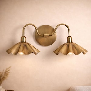 Può includere: Applique da parete color ottone con due luci. Ogni luce ha un braccio curvo e un paralume a balze. L'applique è montata su una piastra rotonda. Le luci sono accese.