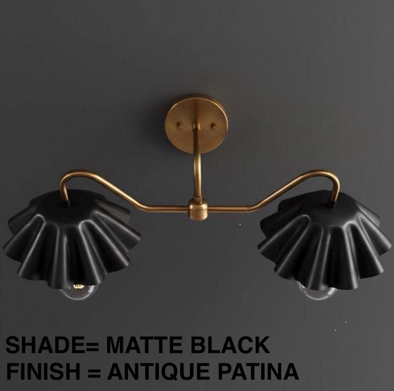 May include: A matte black wall sconce with two lights. The fixture has a brass finish and two ruffled, matte black shades. The text on the image reads: SHADE= MATTE BLACK, FINISH= ANTIQUE PATINA.