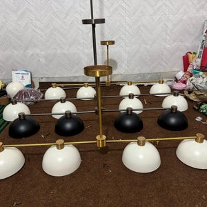 May include: A multi-light chandelier featuring a central gold-toned metal rod with horizontal arms extending outward. The fixture includes numerous dome-shaped shades in matte white and black finishes, each attached to the frame with gold-toned hardware.