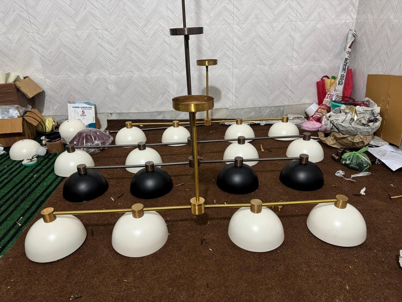 May include: A multi-light chandelier featuring a central gold-toned metal rod with horizontal arms extending outward. The fixture includes numerous dome-shaped shades in matte white and black finishes, each attached to the frame with gold-toned hardware.