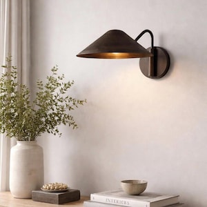 May include: A dark bronze wall sconce with a conical shade and a curved arm. The sconce is mounted on a round backplate. The image also shows a white vase with greenery, a wooden tray with a bowl of nuts, and two books.