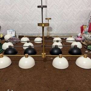 May include: A large multi-light chandelier featuring a central brass-toned rod with horizontal arms. The fixture includes twelve dome-shaped shades in matte black and off-white, each attached to the frame with small brass-toned cylindrical connectors.
