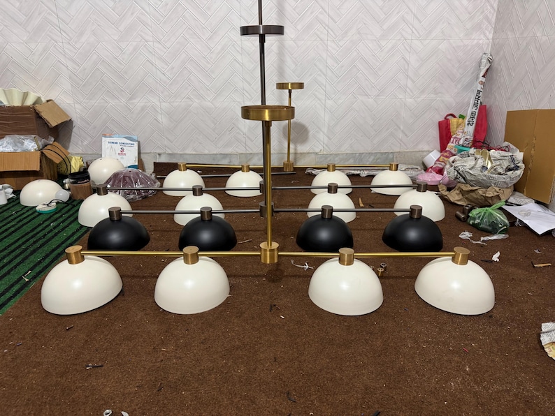 May include: A large multi-light chandelier featuring a central brass-toned rod with horizontal arms. The fixture includes twelve dome-shaped shades in matte black and off-white, each attached to the frame with small brass-toned cylindrical connectors.