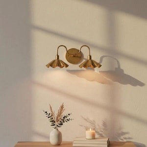 May include: A gold-toned wall sconce with two lights, each featuring a scalloped shade. The sconce is mounted on a cream-colored wall. A small vase with dried flowers, a candle, and books are on a wooden table below.