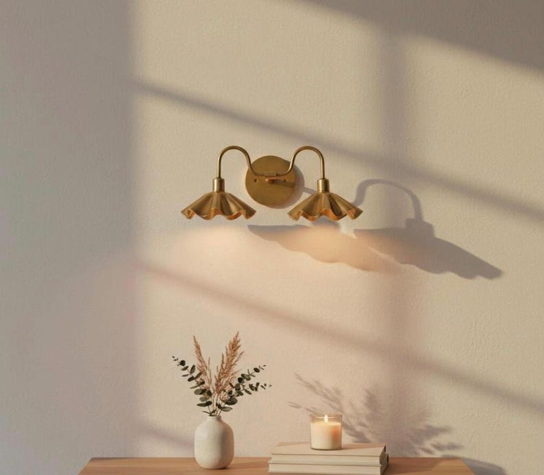 May include: A gold-toned wall sconce with two lights, each featuring a scalloped shade. The sconce is mounted on a cream-colored wall. A small vase with dried flowers, a candle, and books are on a wooden table below.