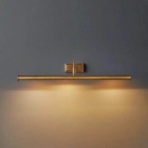 Peut inclure: Une applique murale horizontale en laiton pour tableau, dotée d'une base rectangulaire fixée au mur et d'un long tube cylindrique. Le luminaire émet une lueur chaude dirigée vers le bas, éclairant la surface du mur sous la barre métallique.