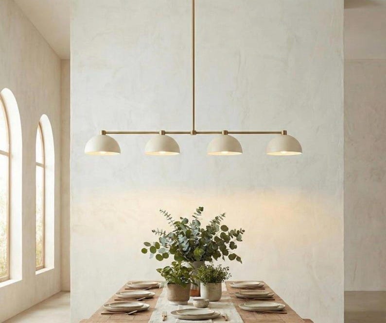 May include: A linear pendant light fixture featuring a long, thin brass-toned metal rod suspended from the ceiling. It holds four evenly spaced, matte off-white dome-shaped shades that direct light downward, creating a clean and symmetrical design.