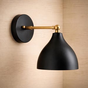 以下が含まれることがあります： ゴールドカラーのアームとアクセントが付いた黒いウォールスコンス。スコンスは丸いベースとベル型のシェードを備えています。この照明器具は、明るい木製のパネル壁に取り付けられています。