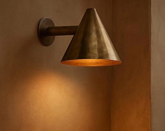 May include: A brass-toned wall sconce with a conical shade. The sconce is mounted on a wall and has a cylindrical arm extending from a circular base. The shade is angled downward, and the interior is illuminated, casting a warm glow.