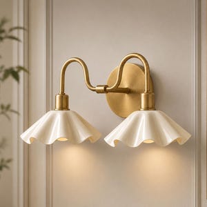 May include: A double wall sconce featuring a brushed brass finish frame with two curved arms extending from a circular backplate. Two off-white, fluted, bell-shaped shades are attached to the ends, directing light downward from the fixture.