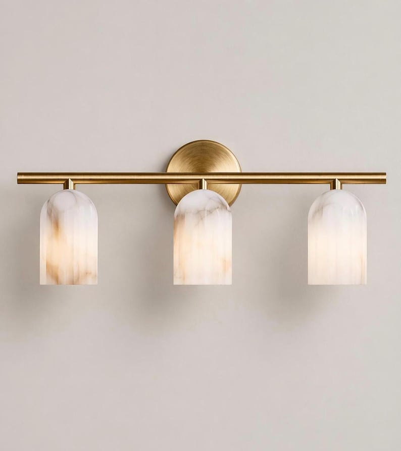 May include: A three-light vanity fixture with a brass finish. The fixture features three arched, white and tan-colored glass shades. The fixture is mounted on a round brass backplate and a horizontal brass bar.