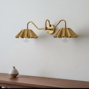 May include: A gold-toned, two-light vanity light fixture with ruffled shades and exposed bulbs. The fixture is mounted on a white wall. A small, two-toned vase sits on a wooden surface below.