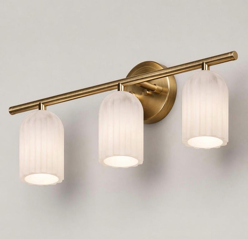 May include: A three-light vanity fixture with a brass finish. The fixture features three frosted glass shades with a ribbed design. The shades are cylindrical with a scalloped bottom edge. The fixture is mounted on a round brass backplate.