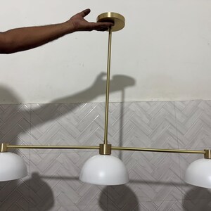 May include: A linear pendant light fixture featuring a horizontal brass-toned metal rod suspended from a matching circular ceiling canopy. The fixture includes three evenly spaced white dome-shaped shades attached to the central horizontal bar.