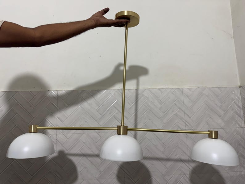 May include: A linear pendant light fixture featuring a horizontal brass-toned metal rod suspended from a matching circular ceiling canopy. The fixture includes three evenly spaced white dome-shaped shades attached to the central horizontal bar.