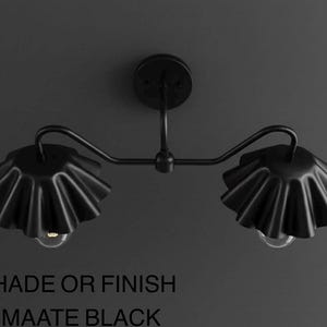May include: Matte black two-light wall sconce. The fixture features two ruffled shades and a curved arm design. The text at the bottom of the image reads "SHADE OR FINISH = MAATE BLACK".