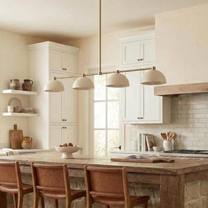 May include: A linear pendant light fixture featuring a long, thin brass-toned metal rod suspended from the ceiling. It holds four evenly spaced, dome-shaped shades in a matte off-white finish, directed downward to provide focused illumination.