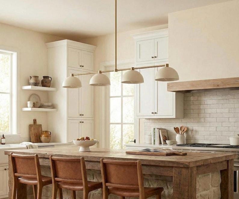 May include: A linear pendant light fixture featuring a long, thin brass-toned metal rod suspended from the ceiling. It holds four evenly spaced, dome-shaped shades in a matte off-white finish, directed downward to provide focused illumination.