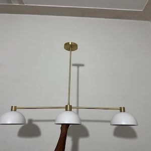 May include: A linear chandelier featuring a central brass-toned rod and horizontal bar supporting three white dome-shaped shades. The fixture includes a circular brass-toned ceiling canopy and matching hardware connecting the shades to the horizontal bar.