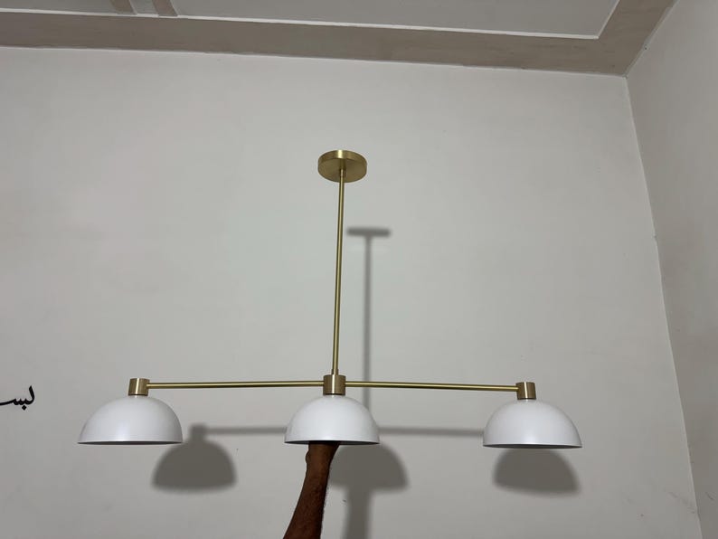 May include: A linear chandelier featuring a central brass-toned rod and horizontal bar supporting three white dome-shaped shades. The fixture includes a circular brass-toned ceiling canopy and matching hardware connecting the shades to the horizontal bar.