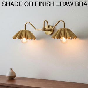 May include: A raw brass wall sconce with two lights, each with a scalloped shade. The sconce is mounted on a white wall. The text at the top reads "SHADE OR FINISH = RAW BRASS".