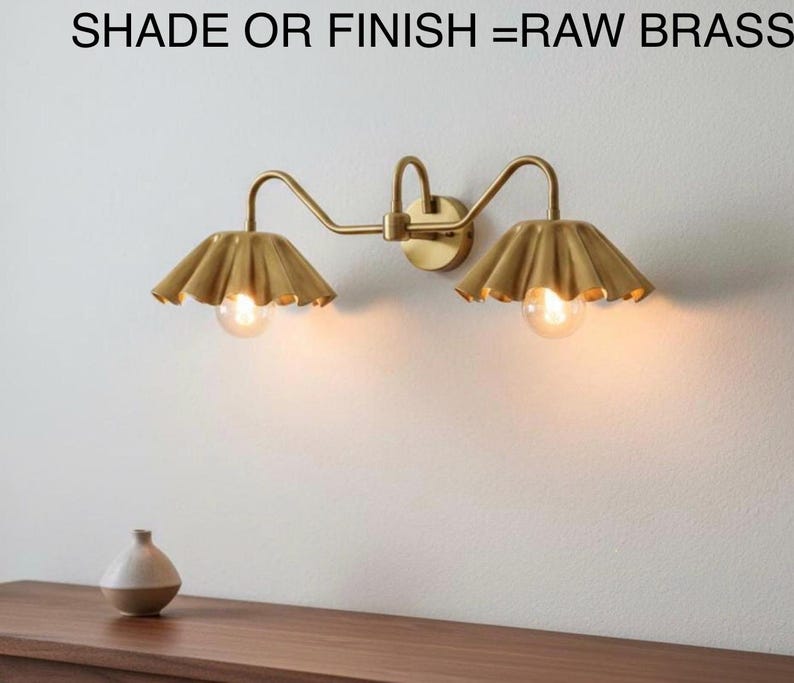 May include: A raw brass wall sconce with two lights, each with a scalloped shade. The sconce is mounted on a white wall. The text at the top reads "SHADE OR FINISH = RAW BRASS".