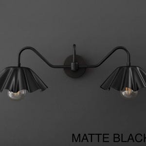 May include: Matte black wall sconce with two lights. Each light has a ruffled shade and a clear bulb. The fixture is mounted on a dark gray wall. The text "MATTE BLACK" is at the bottom.
