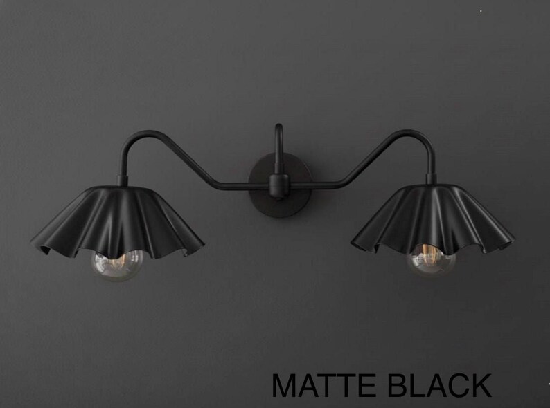 May include: Matte black wall sconce with two lights. Each light has a ruffled shade and a clear bulb. The fixture is mounted on a dark gray wall. The text "MATTE BLACK" is at the bottom.