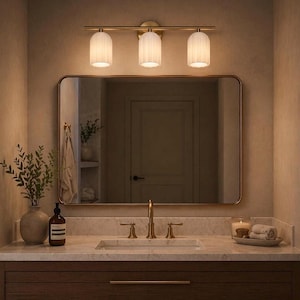 May include: A bathroom vanity with a rectangular mirror, a three-light fixture, and a wooden cabinet. The light fixture has a gold finish and three white, cylindrical shades. A vase of greenery and a soap dispenser are on the countertop.