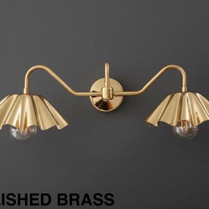 May include: A polished brass wall sconce with two lights. Each light has a scalloped shade and a clear bulb. The fixture is mounted on a dark gray wall. The text "POLISHED BRASS" is at the bottom.