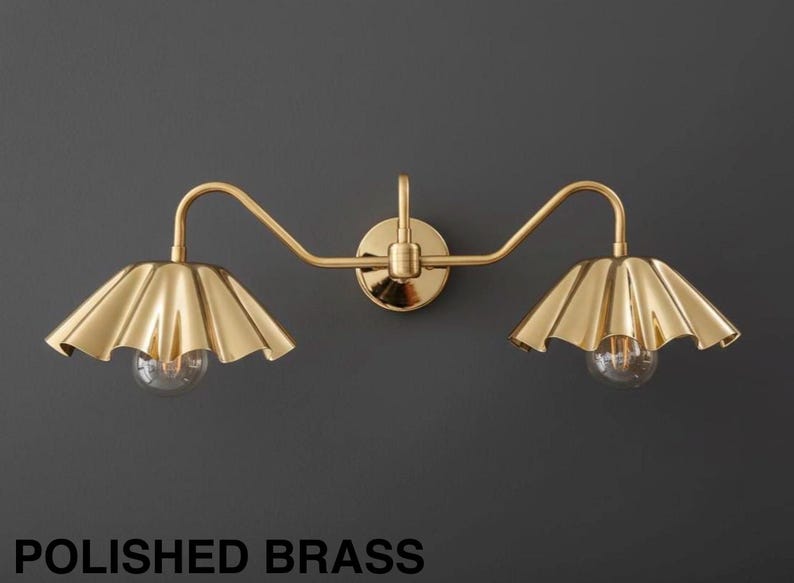 May include: A polished brass wall sconce with two lights. Each light has a scalloped shade and a clear bulb. The fixture is mounted on a dark gray wall. The text "POLISHED BRASS" is at the bottom.