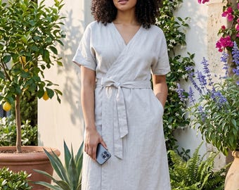 May include: A light gray wrap dress with short sleeves, a V-neckline, and a matching fabric tie at the waist. The dress features a knee-length hem and is worn with brown strappy sandals. The garment has a simple, clean design with visible side pockets.