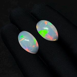 Könnte beinhalten: Zwei birnenförmige Cabochon-Opale mit einer durchscheinenden weißen Basis und lebhaften Blitzen in Neongrün und hellem Orange. Die Edelsteine sind glatt, poliert und zeigen einen ausgeprägten irisierenden Glanz auf ihren Oberflächen.