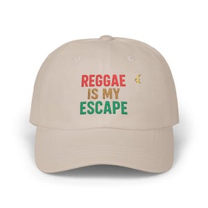 May include: A beige baseball cap with the text "REGGAE IS MY ESCAPE" in red, green, and brown. A small yellow and green graphic is to the right of the text. The cap has a curved brim and a fabric strap closure.