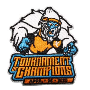 Può includere: Toppa ricamata raffigurante una creatura simile a uno yeti con la pelle blu, una fascia arancione e la bocca aperta. La toppa recita "TOURNAMENT OF CHAMPIONS" in arancione e blu, con "APRIL 26 2025" sotto. Il disegno è delineato in nero.