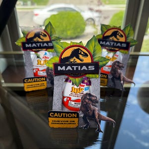 May include: Three Kinder Joy eggs with custom dinosaur-themed decorations. Each egg is adorned with a dinosaur silhouette, the name "MATIAS", and a "CAUTION" sign in Spanish. The decorations feature a Jurassic Park aesthetic.