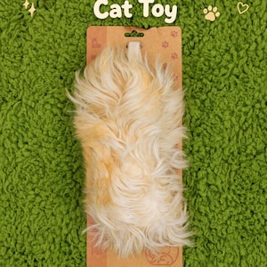 Puede incluir: Juguete para gatos de pelo de cabra cremoso y naranja claro, fijado a un respaldo de cartón marrón. El texto "Creamy Goat Fluff Cat Toy" está en letras blancas sobre el juguete. El fondo es verde.