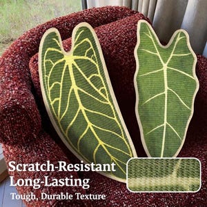 May include: Two green leaf-shaped decorative pillows with yellow vein details and a cream-colored border. The pillows are on a red textured surface. The image also includes a close-up of the fabric texture and the text "Scratch-Resistant Long-Lasting Tough, Durable Texture."