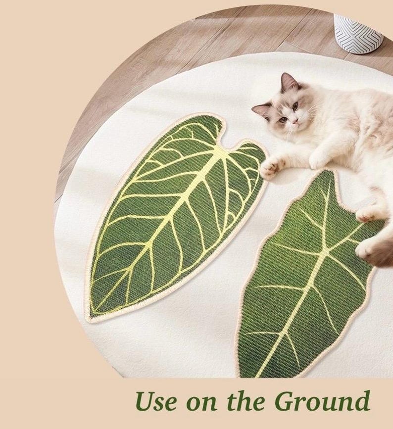 May include: A white round rug with two green leaf-shaped mats. The mats have a detailed vein design in yellow. A fluffy cat is lying on the rug. The text "Use on the Ground" is at the bottom.