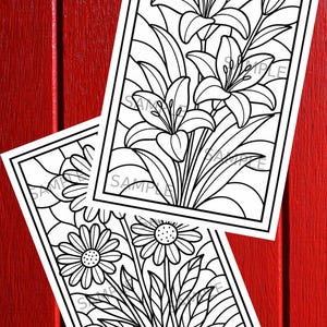 May include: Two black and white floral coloring pages featuring lilies and daisies. The images are in a rectangular frame with the word "SAMPLE" printed on them. The background is a red wooden surface.