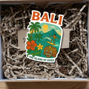 Può includere: Un adesivo di Bali con un design colorato con un surfista, palme e una tradizionale maschera balinese. L'adesivo è in una scatola con carta sminuzzata. Il testo sull'adesivo dice "BALI" e "THE ISLAND OF GODS". La scatola ha anche il testo "DIGITAL DOWNLOAD" e "Bali Sticker".