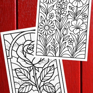 May include: Two black and white floral coloring pages. One features a rose in a stained glass window design. The other has a variety of flowers and foliage. The pages are on a red wooden surface.