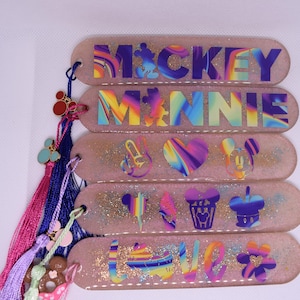 Puede incluir: Una colección de cinco marcapáginas brillantes de color rosa claro con diseños y texto coloridos. El marcapáginas superior dice "MICKEY" y el segundo "MINNIE". Cada marcapáginas tiene una borla con abalorios.