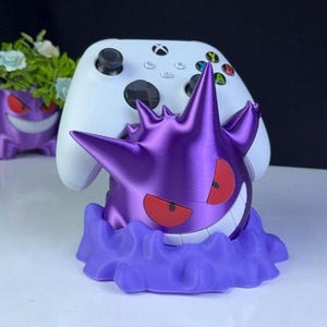 Może przedstawiać: Fioletowy stojak na kontroler w stylu Gengar z białym kontrolerem Xbox. Stojak ma uśmiechniętą twarz Gengara z czerwonymi oczami i podstawą w kształcie chmury. W tle widoczna jest podobna fioletowa doniczka.