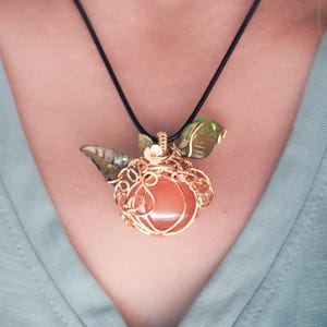 May include: A pendant featuring a round, translucent orange stone encased in intricate copper wire wrapping, accented with two green glass leaves and a small flower, suspended from a simple black cord necklace against a light green shirt.